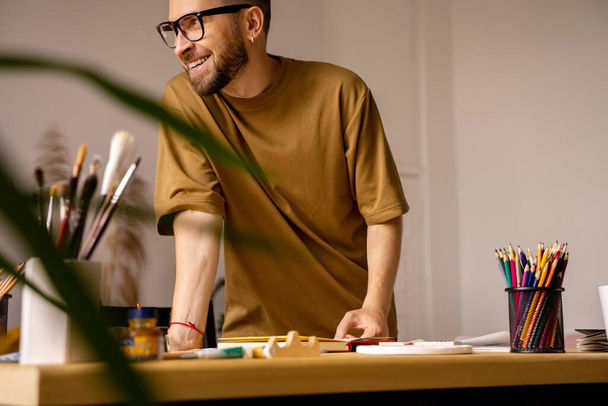ワークショップで机の上に立っている若いアーティスト. アーティストのスタジオで働くインスピレーションを見つけるプロセス. 幸せな男が微笑んでいる. ライフスタイル。 リアルな感情 - 写真・画像