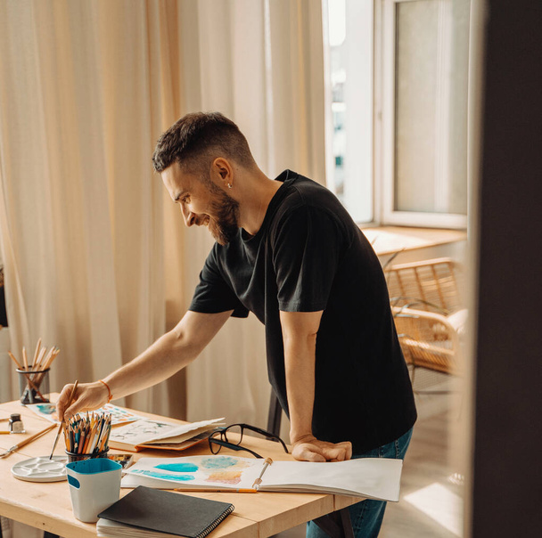 Um jovem artista feliz, sorrindo, faz um esboço com um pincel no estúdio. Uma pessoa criativa está em pé sobre um desktop com um pincel na mão.Estúdio, estilo de vida, processo de criação de uma obra de arte. - Foto, Imagem