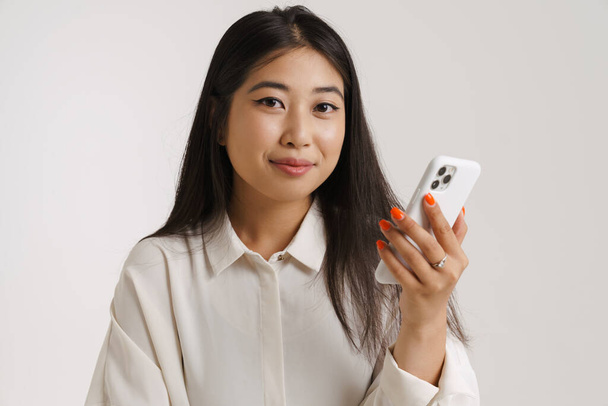 Giovane donna asiatica sorridente e utilizzando il telefono cellulare isolato su parete bianca - Foto, immagini