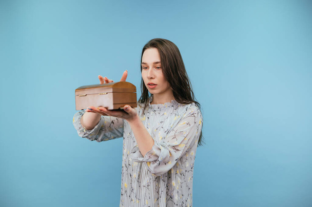 Cute lady in dress opens a wooden box with jewelry on a blue background with a serious face on a blue background. - Photo, Image
