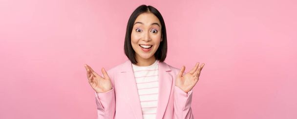 Retrato de una mujer de negocios asiática mirando sorprendida a la cámara, aplaudiendo y mirando emocionada, sonriendo, posando sobre fondo rosa - Foto, Imagen