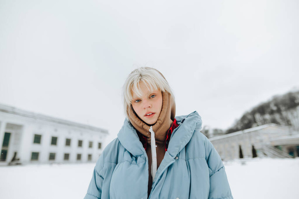 Portret van een jong meisje met blond haar en warme casual kleding staat op een besneeuwde straat en kijkt naar de camera. - Foto, afbeelding