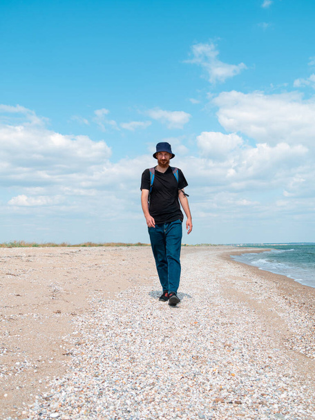 Smiling bearded millennial man in bucket hat black t-shirt walking on empty beach. Authentic male tourist lifestyle photo. Hipster guy outdoor. Solo travel adventure concept Active walking backpacking - Fotografie, Obrázek