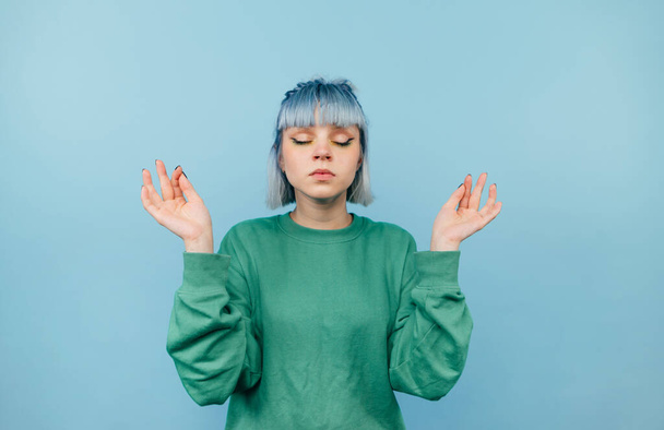 Серьезная девушка в зеленой толстовке стоит на синем фоне и медитирует с закрытыми глазами. Isolated - Фото, изображение