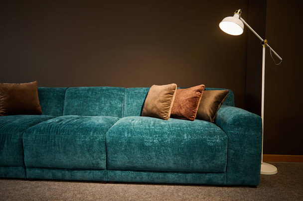 Exposición de muebles tapizados con estilo moderno en la sala de exposición de una tienda de muebles. Concéntrate en un sofá de terciopelo suave turquesa y almohadas marrones iluminadas por una lámpara sobre un fondo de pared marrón - Foto, imagen