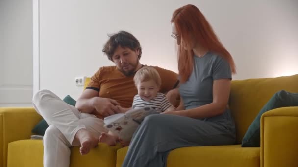 Perhe istuu keltaisella sohvalla ja opettaa vauvaa lukemaan vauvan piirustus kirjaan - Materiaali, video
