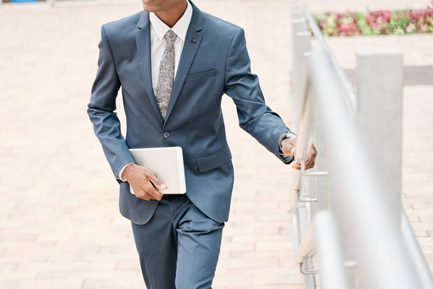 nierozpoznawalny człowiek przemierzający miasto. młody biznesmen w garniturze i krawacie z cyfrowym tabletem w ręku. - Zdjęcie, obraz