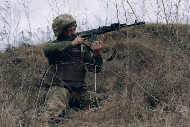 ウクライナの兵士は、乾燥した草で丘の背景にロシアの敵にkalashnikovアサルトライフルから座っていると目的。ウクライナにおけるロシアの軍事侵攻の概念。ウクライナとヨーロッパでの戦争. - 写真・画像