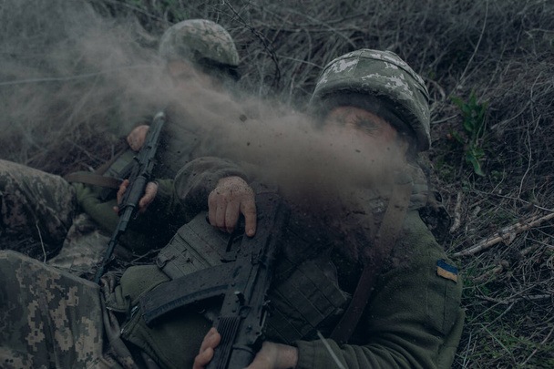 Dva ukrajinští vojáci leží s kalašnikovovými útočnými puškami na suché trávě během dělostřeleckého bombardování ruským nepřítelem. Koncept ruské vojenské invaze na Ukrajině. Válka na Ukrajině a v Evropě. - Fotografie, Obrázek