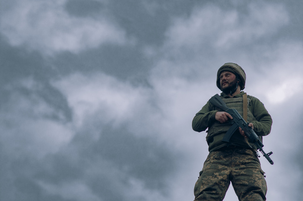 ウクライナ兵は空と雲を背景にカラシニコフのアサルトライフルで立っている。ウクライナにおけるロシアの軍事侵攻の概念。ウクライナとヨーロッパでの戦争。コピースペース付き画像. - 写真・画像