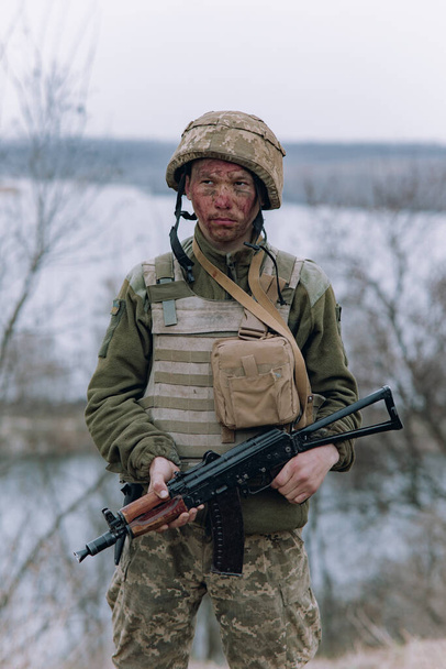 Ουκρανός στρατιώτης στέκεται με kalashnikov τουφέκι επίθεση στα χέρια του σε φόντο του ουρανού και του ποταμού. Έννοια της ρωσικής στρατιωτικής εισβολής στην Ουκρανία. Πόλεμος στην Ουκρανία και την Ευρώπη. - Φωτογραφία, εικόνα