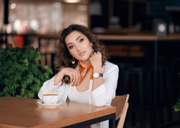 Деловая молодая женщина сидит за столом в кафе, пьет кофе, на ней белая куртка - Фото, изображение