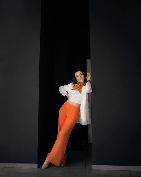 Eine junge Geschäftsfrau steht an einer schwarzen Wand und trägt eine weiße Jacke und eine orangefarbene Hose - Foto, Bild