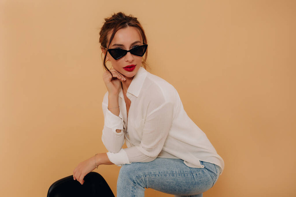 Portrait of european dark-haired girl in dark glasses and white shirt with red lips is posing during photoshoot on beige background with place for text - Fotó, kép