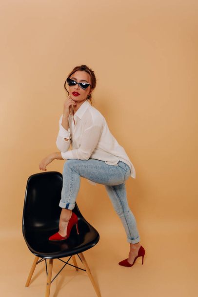 Full-lenght portrait of spectacular lovely woman with dark hair is wearing white shirt and jeans in dark glasses with red lips is raised up one leg on chair. Lovely woman is posing over isolated wall - Photo, image