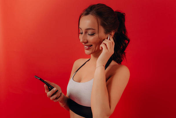 Excited lovely girl with wavy dark hair is wearing sport top is listening music in headphones and holding smartphone before training. Smiling girl preparing for training over red background - Zdjęcie, obraz