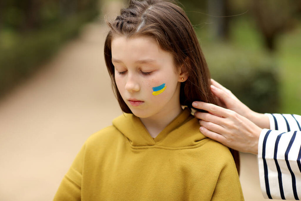 Közelkép a gondoskodó fiatal anya fonat haj szomorú kislány zászlóval az arcon a szabadban. Béke, háború nélkül, segítség, az orosz agresszió megállítása. Oroszország lerohanása Ukrajnában. Kiváló minőségű fénykép.. - Fotó, kép