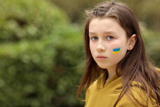 the face of a frightened girl, painted on her cheek in the yellow-blue colors of the Ukrainian flag, a request for help. Children ask for peace. High quality photo. High quality photo - Foto, Imagem