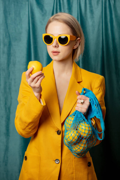 Sarı güneş gözlüklü ve ceketli şık Ukraynalı kadın yeşil perde arkasında ağ torbasında limonlar - Fotoğraf, Görsel