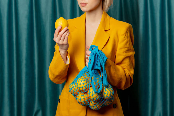 Stylish ukrainian woman in yellow sunglasses and jacket with lemons in net bag on green curtains background - Photo, Image