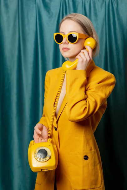 Elegante mujer ucraniana en gafas de sol amarillas y chaqueta con teléfono de reparto sobre fondo de cortinas verdes - Foto, Imagen