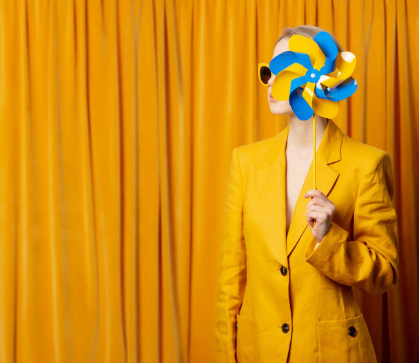 Stijlvolle Oekraïense vrouw in gele zonnebril en jas met pinwheel op gordijnen achtergrond - Foto, afbeelding