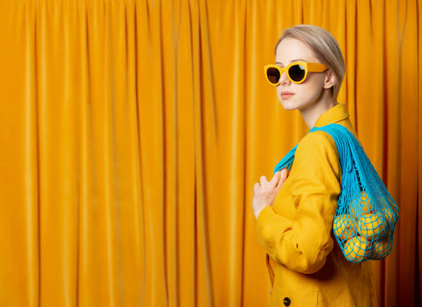 Stylish ukrainian woman in yellow sunglasses and jacket with lemons in net bag on curtains background - Foto, afbeelding