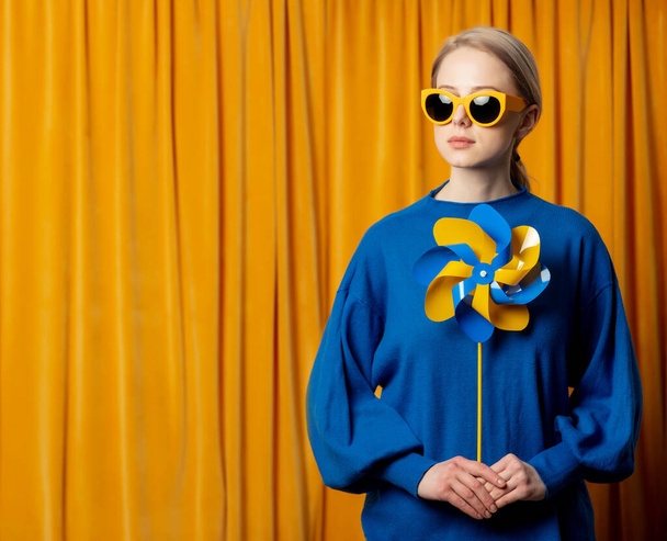 Stylová ukrajinská žena ve žlutých slunečních brýlích a modrém svetru s větrníkem v ukrajinských vlajkových barvách na pozadí záclony - Fotografie, Obrázek