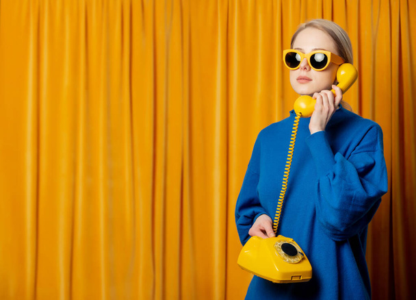 Elegante mujer ucraniana en gafas de sol amarillas y suéter azul con teléfono de línea sobre fondo de cortinas - Foto, imagen
