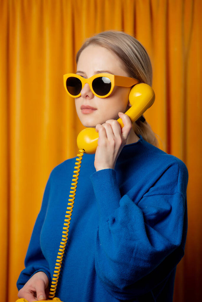 Stilvolle ukrainische Frau in gelber Sonnenbrille und blauem Pullover mit Wählscheibe Telefon auf Vorhang Hintergrund - Foto, Bild