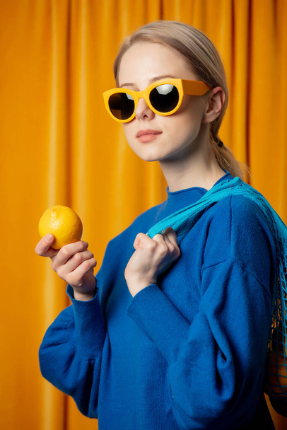 Stylish ukrainian woman in yellow sunglasses and blue sweater with lemons in net bag on curtains background - Foto, afbeelding
