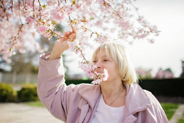 Senior woman in pink coat smells blooming branch of sakura during her walking in the spring garden - Photo, Image