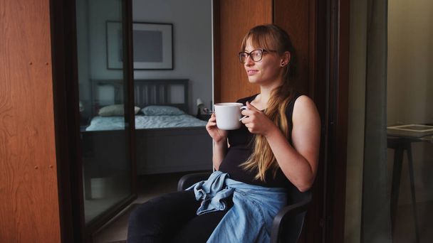 Πορτρέτο της νεαρής καυκάσιας κοκκινομάλλας που απολαμβάνει ένα μεγάλο φλιτζάνι καφέ ή τσάι στο μπαλκόνι της στο σπίτι.  - Φωτογραφία, εικόνα