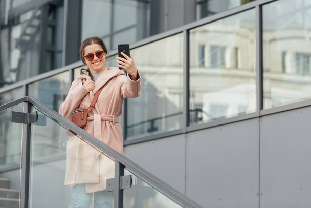 Woman in pink dress take selfie on smarthone on grey urban background outdoor - Foto, imagen