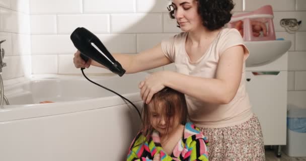 Gelukkige moeder droogt haar dochters haar. Avondritueel van moeder en dochter in de badkamer. Gezin in huiskleding. - Video