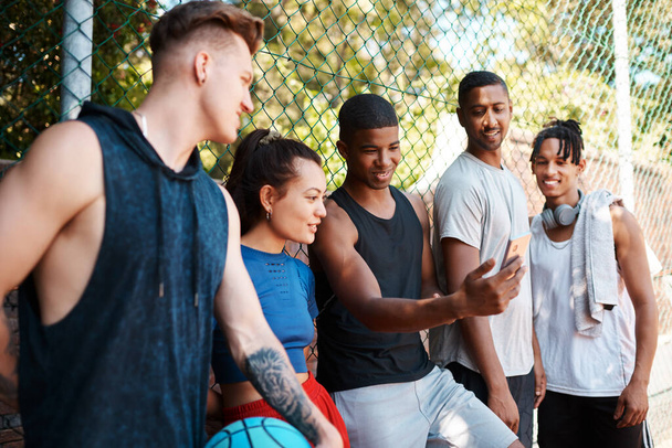 Das muss man sehen. Aufnahme einer Gruppe sportlicher junger Leute, die sich etwas auf einem Handy anschauen, während sie im Freien an einem Zaun stehen. - Foto, Bild