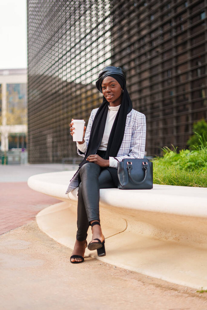 Stilvolle ernste schwarze Frau mit Kopftuch beim Kaffee auf Bank - Foto, Bild