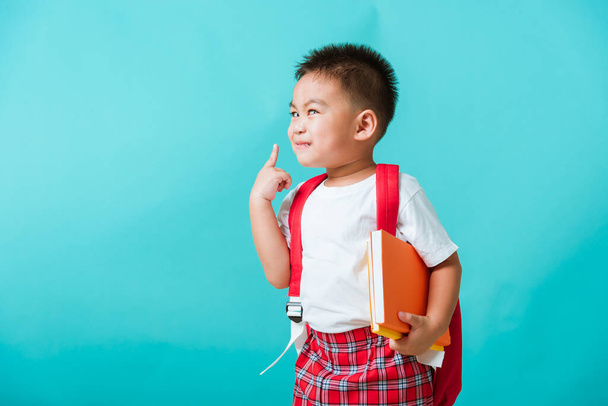 Back to school concept. Portrait Asian happy funny cute little child boy smile hug books and point finger to side away space, isolated blue background. Kid from preschool kindergarten with school bag - Foto, afbeelding
