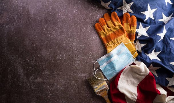 Концепция "Счастливого дня труда во время коронавирусной болезни" (Ковид-19). Американский флаг с различными строительными инструментами и медицинской маской на фоне темного камня. - Фото, изображение