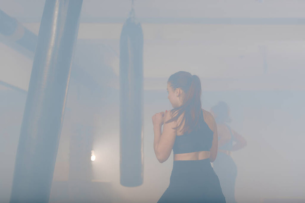 Caucasian woman in sportswear with a ponytail, kicking the boxing bag inside a gym with a smokey background - Foto, Imagen