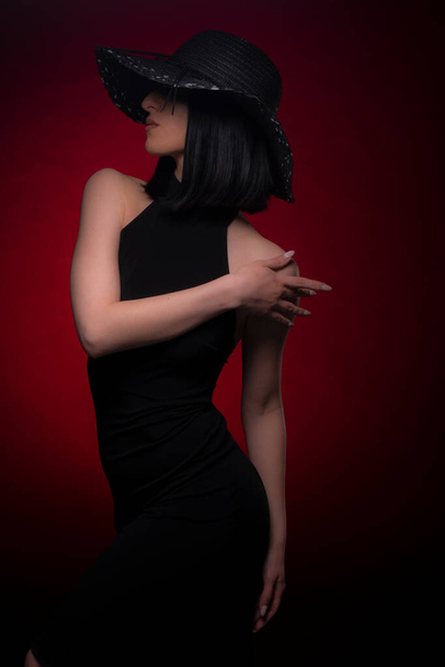 Κομψό και όμορφο κορίτσι ποζάρει στο σύνολο, ενώ αγγίζει τον ώμο της σε κόκκινο φόντο στο μαύρο φόρεμα της - Φωτογραφία, εικόνα