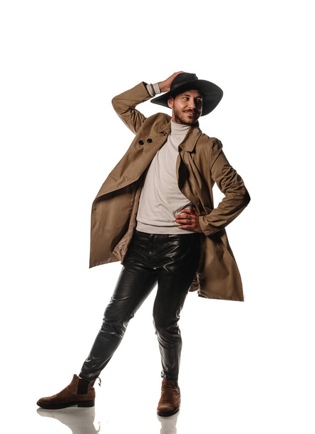 Cool young man dressed in beige pullover and long coat posing in modern studio against white background - Photo, Image