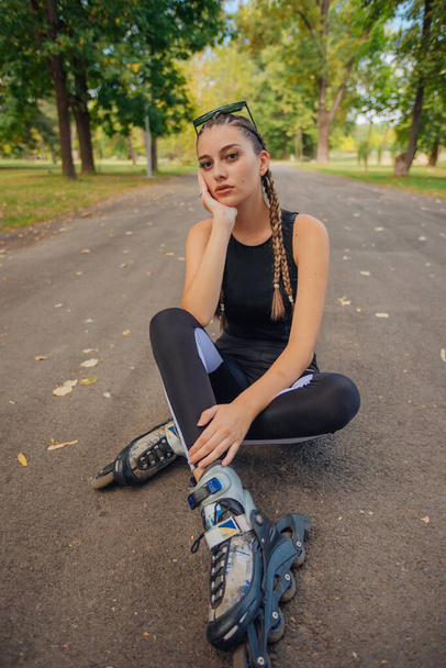 Удивительная молодая женщина с косичками, позирующая на роликовых коньках, сидя на дороге в парке - Фото, изображение