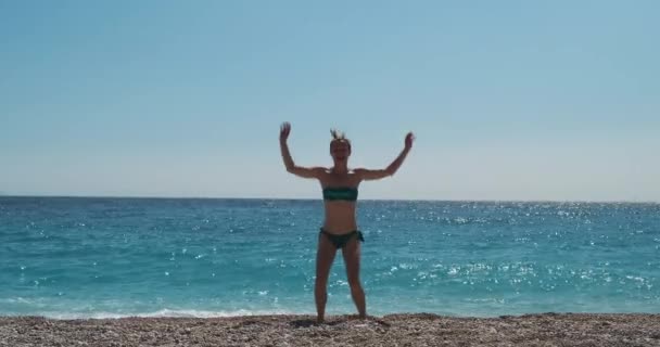 Una hermosa chica en bikini salta a una playa de guijarros sobre el fondo del mar azul. Levanta las manos, despreocupada y feliz de vacaciones. Durante el día, tiro ancho - Metraje, vídeo