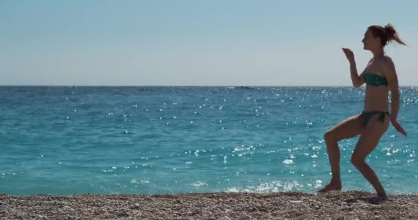 bella ragazza caucasica in costume da bagno in bikini è scherzare da solo sulla spiaggia, sullo sfondo di un perfetto mare turchese. Ballando come un egiziano, braccia ai lati. Di giorno, inquadratura ampia - Filmati, video