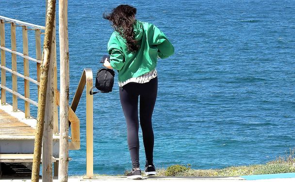 Μελαχρινή σε καλσόν με πράσινη μπλούζα που τραβά φωτογραφίες από τον ωκεανό - Φωτογραφία, εικόνα