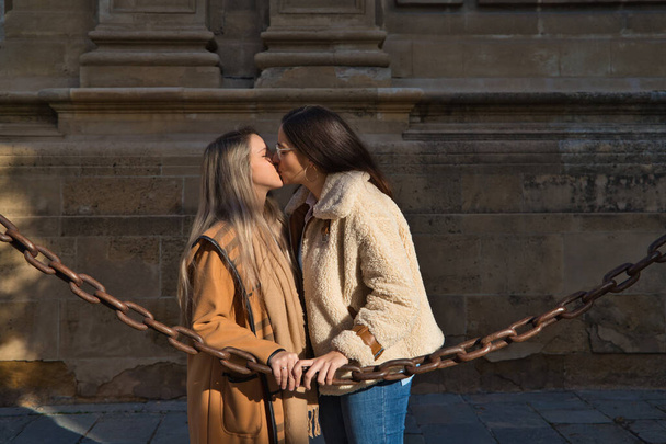 Lesbický pár opírající se o řetěz obklopující gotickou katedrálu. Ženy se líbají v lásce a vášnivě. Lesbický a gay manželský koncept. - Fotografie, Obrázek