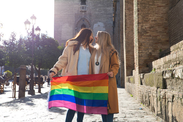 Брак лесбиянок на отдыхе и туризме в Севилье. Они стоят перед собором и во время поцелуя держат в руках флаг гей-гордости. Концепция равенства и прав lgtb. - Фото, изображение