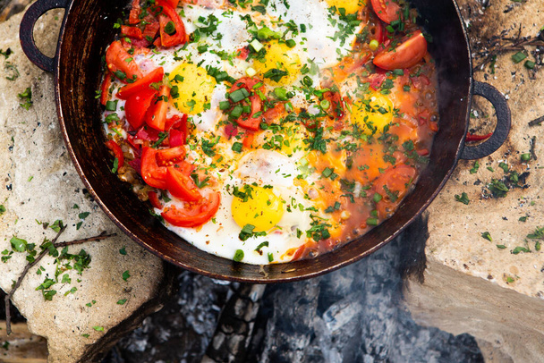 Eggs prepared with vegetables on a frying pan on the fire in the forest. Cooking scrambled eggs over an open fire. Breakfast in nature. Tourist food. - Photo, Image