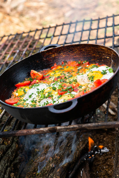 森の中の火の上にフライパンに野菜を入れた卵。火の上でスクランブルエッグを調理する。自然の中で朝食。観光食品. - 写真・画像